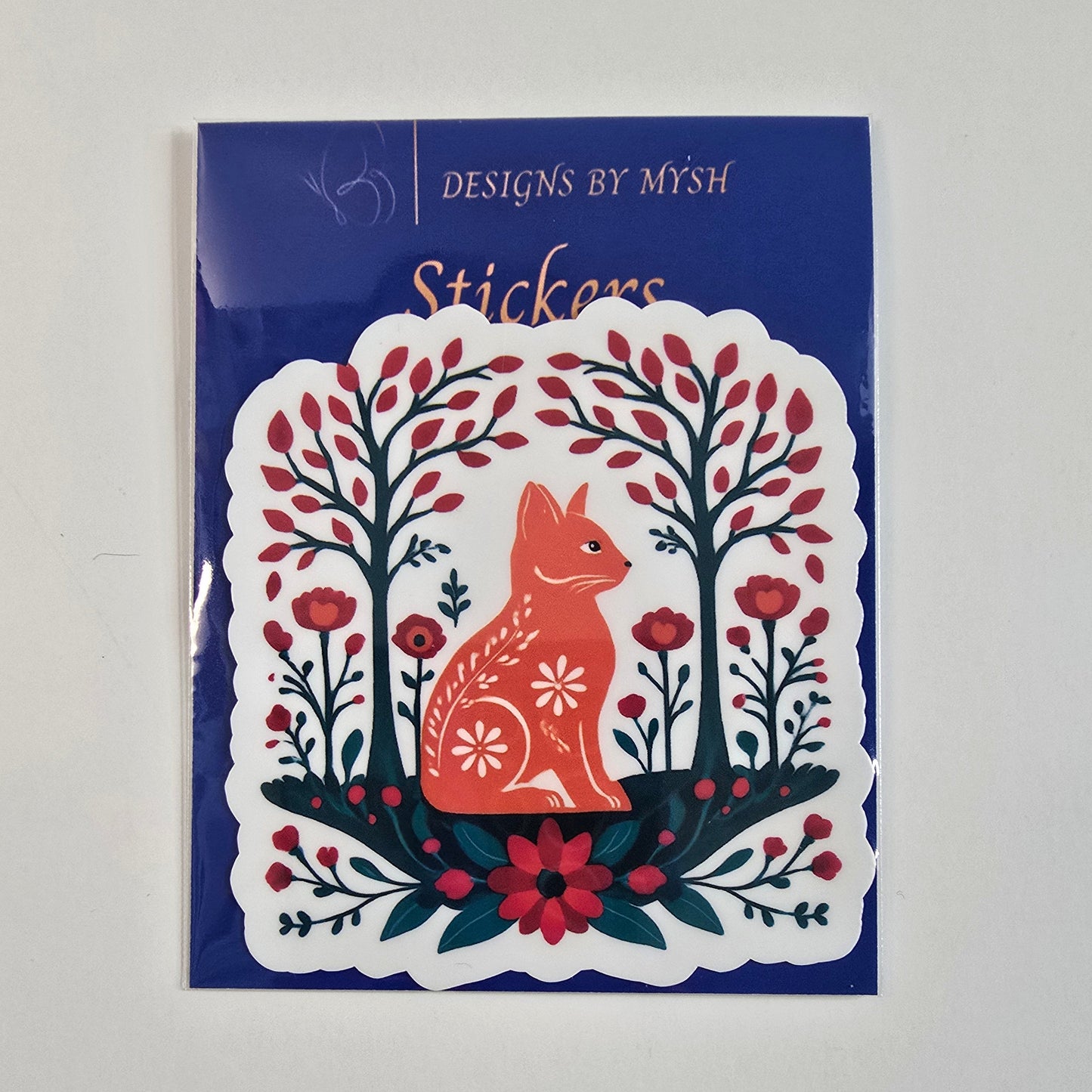 Die-cut vinyl sticker featuring a red folk art-style fox surrounded by symmetrical trees, red flowers, and greenery, mounted on a branded card.