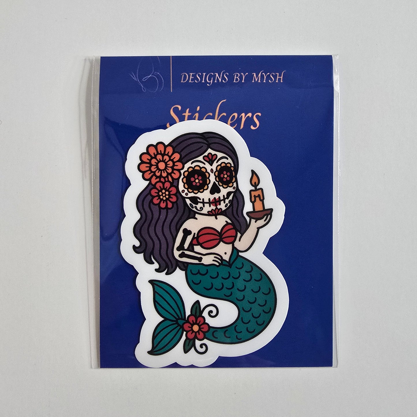 Vinyl sticker of a sugar skull mermaid holding a candle with teal tail, purple hair, and floral details, on branded backing card.