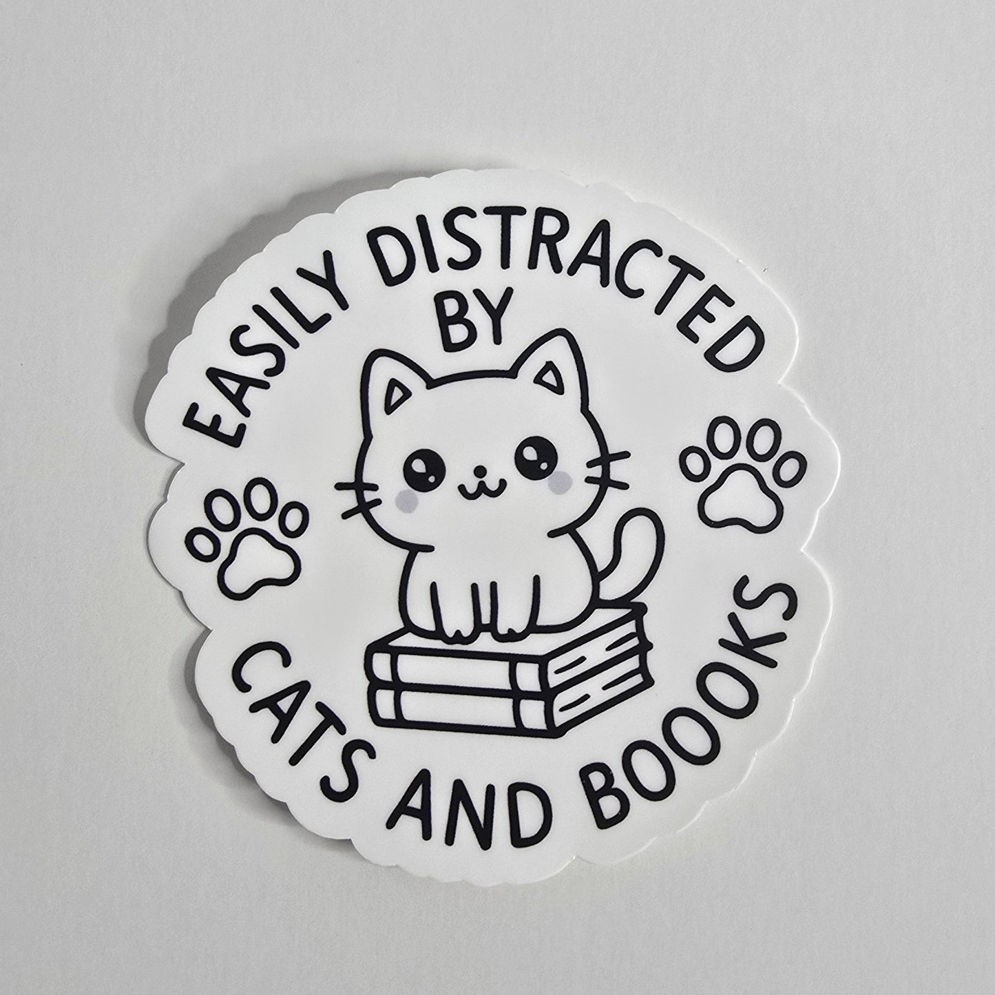 "Die-cut vinyl sticker with a black-and-white kawaii cat sitting on books, surrounded by the text 'Easily Distracted by Cats and Boooks' and two paw prints."