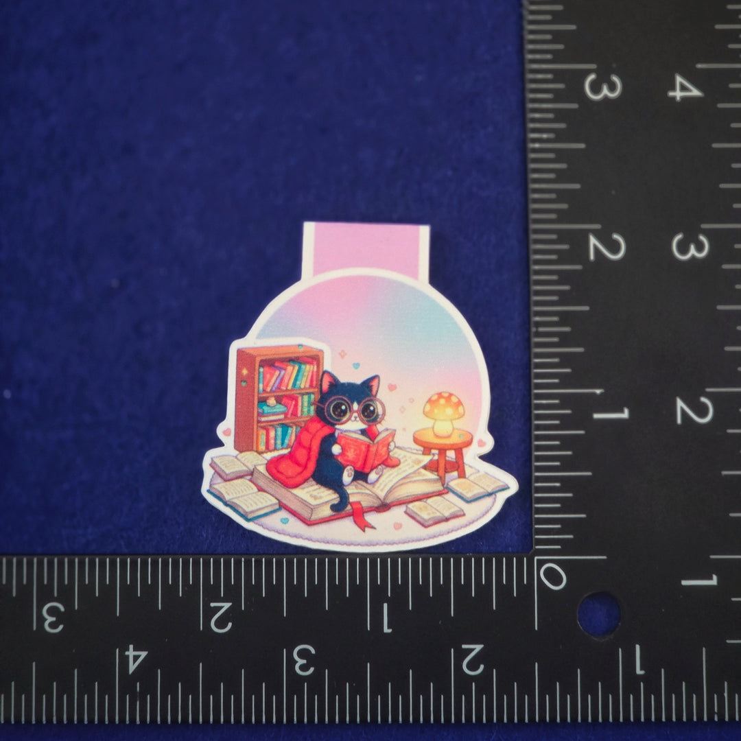 Sticker of a cat in a snow globe with books and a cake, next to a ruler for scale on a dark blue background.