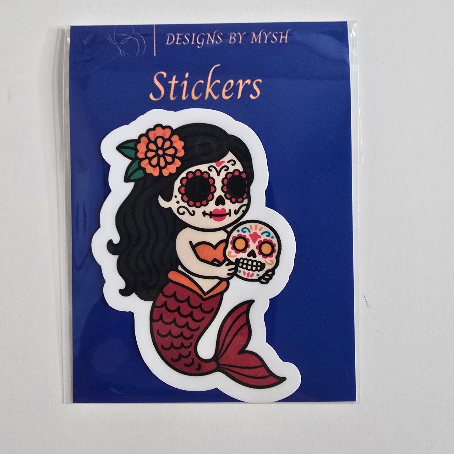 Vinyl sticker of a Day of the Dead mermaid holding a colorful sugar skull, in a cartoon style with red tail, orange top, and floral headpiece.