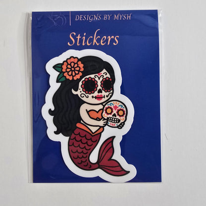 Vinyl sticker of a Day of the Dead mermaid holding a colorful sugar skull, in a cartoon style with red tail, orange top, and floral headpiece.