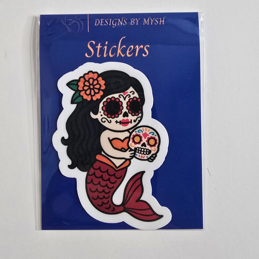 Vinyl sticker of a Day of the Dead mermaid holding a colorful sugar skull, in a cartoon style with red tail, orange top, and floral headpiece.