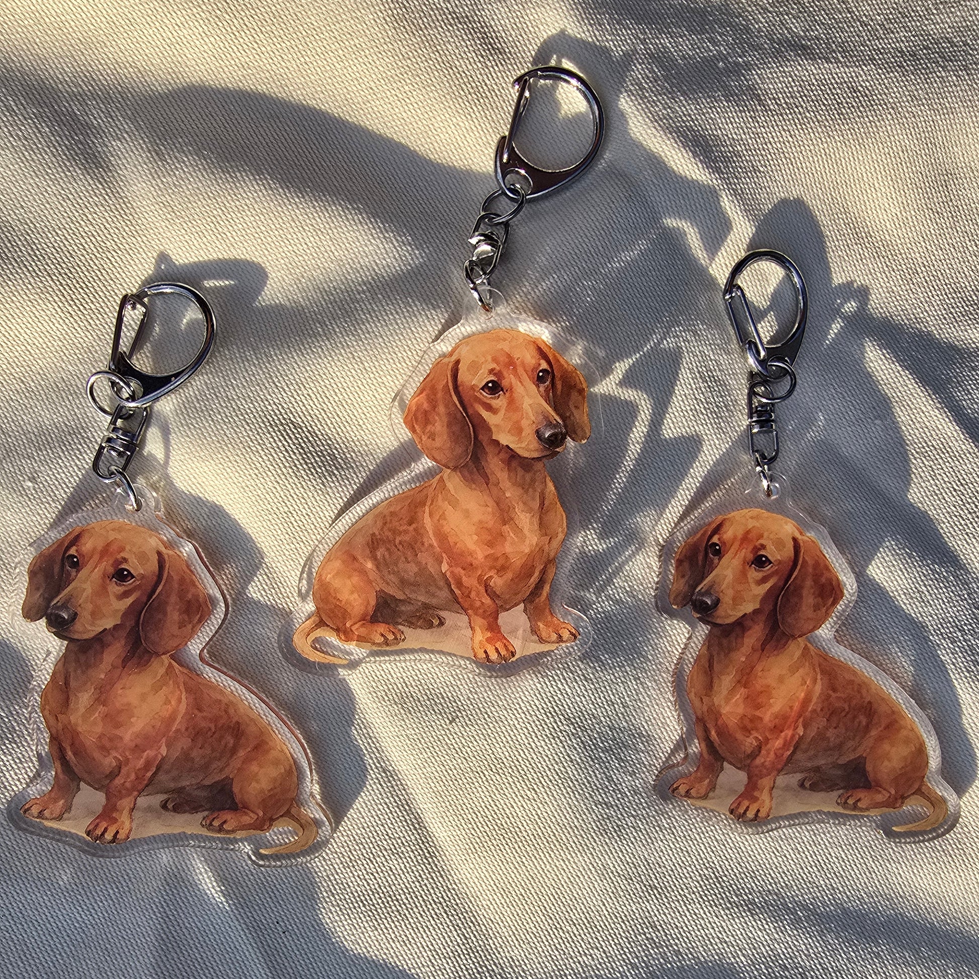 Three red dachshund watercolor acrylic keychains arranged on white fabric in natural light