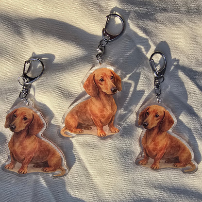 Three red dachshund watercolor acrylic keychains arranged on white fabric in natural light