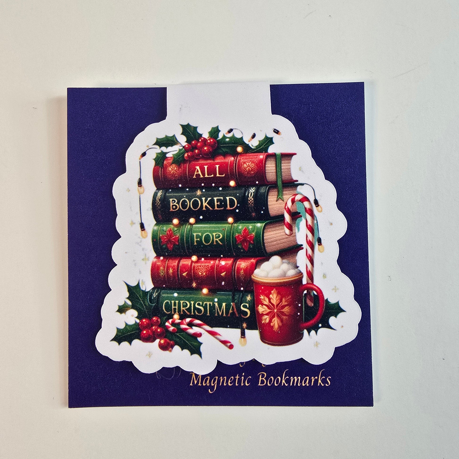 Magnetic bookmark with a stack of Christmas-themed books reading "All Booked for Christmas," with holly, candy cane, and a cocoa mug – by Designs by Mysh.