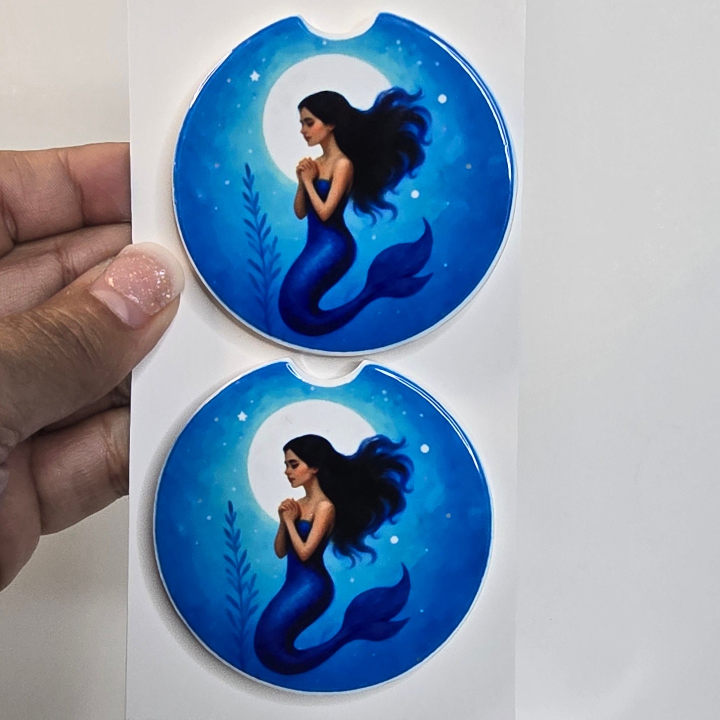 Two blue mermaid car coasters held by a hand on a white background.