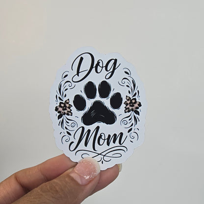 Hand holding a round sticker with 'Dog Mom' text and paw print design against a light background.