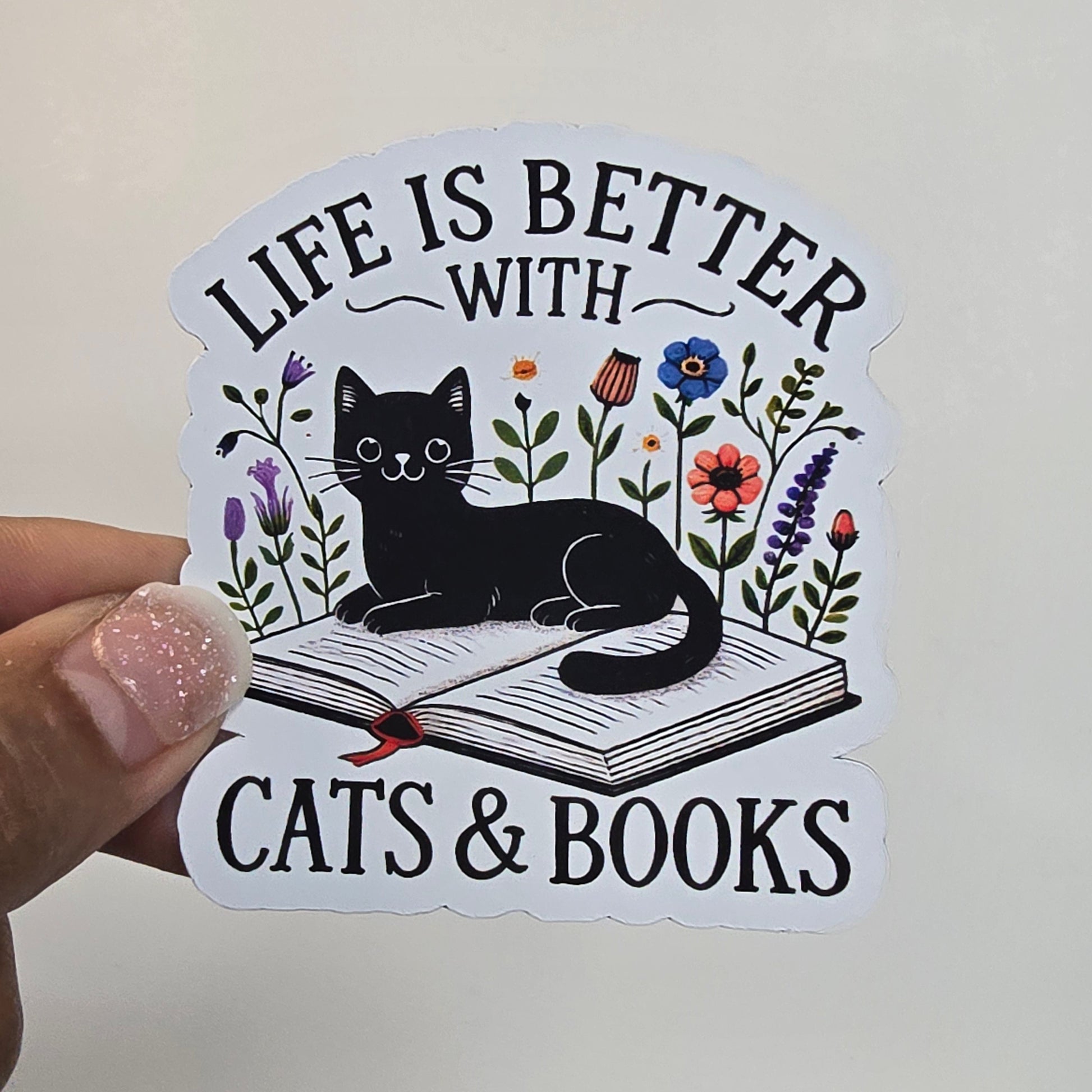 Sticker with a cat, book, and floral design held by a hand against a grid background