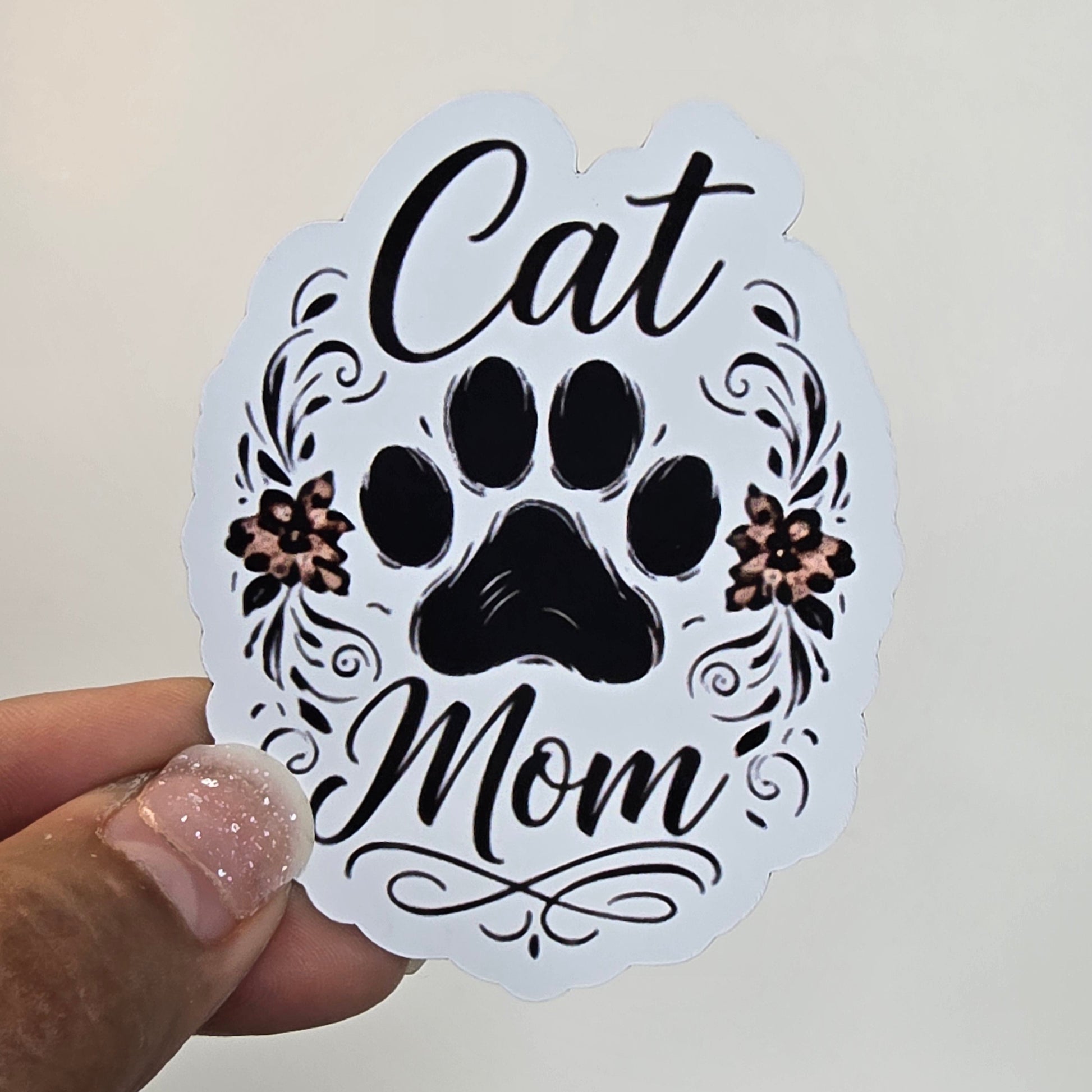 Sticker with 'Cat Mom' text and paw prints held by a hand against a white background.