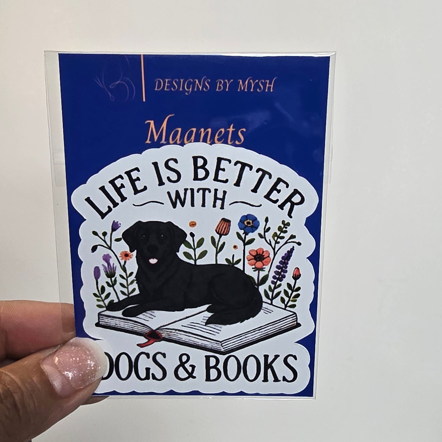 Sticker with 'Life is better with dogs & books' design held by a hand against a white wall.