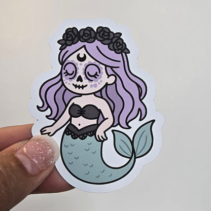 Sticker of a mermaid with purple hair held by a hand against a light background