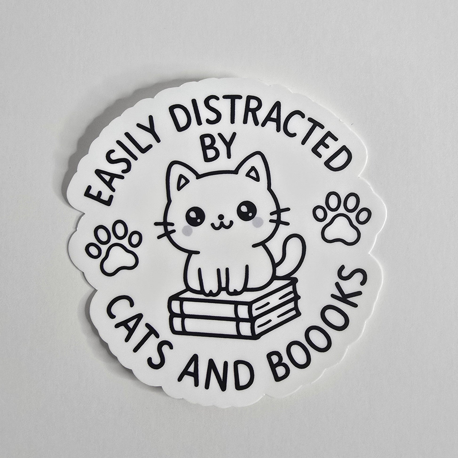 "Die-cut vinyl sticker with a black-and-white kawaii cat sitting on books, surrounded by the text 'Easily Distracted by Cats and Boooks' and two paw prints."