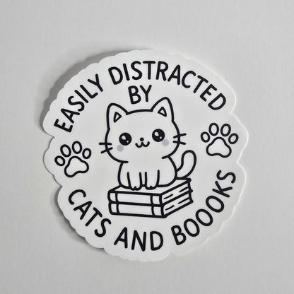 "Die-cut vinyl sticker with a black-and-white kawaii cat sitting on books, surrounded by the text 'Easily Distracted by Cats and Boooks' and two paw prints."