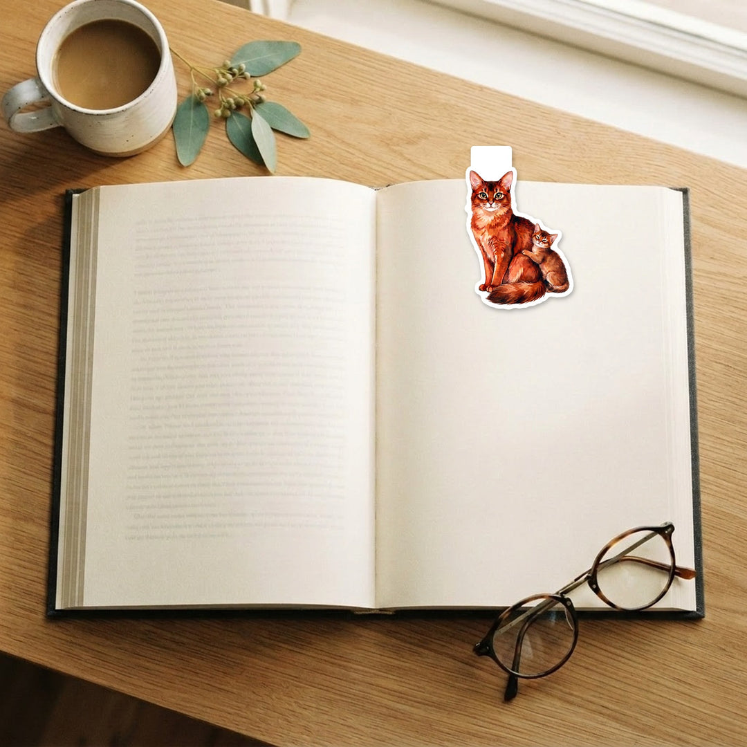 Somali cat and kitten magnetic bookmark on an open book on a wooden desk with coffee and reading glasses