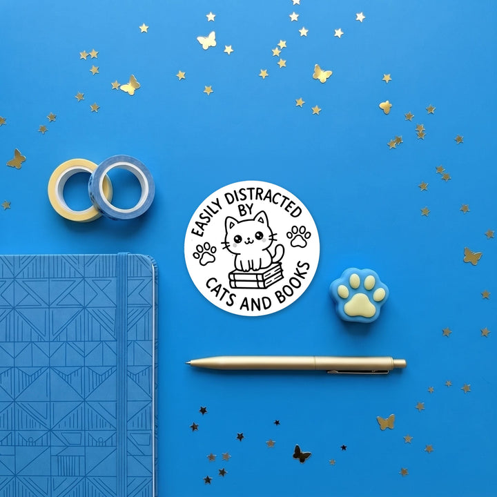 Easily Distracted by Cats and Books sticker in a brand flat lay with a notebook and pen