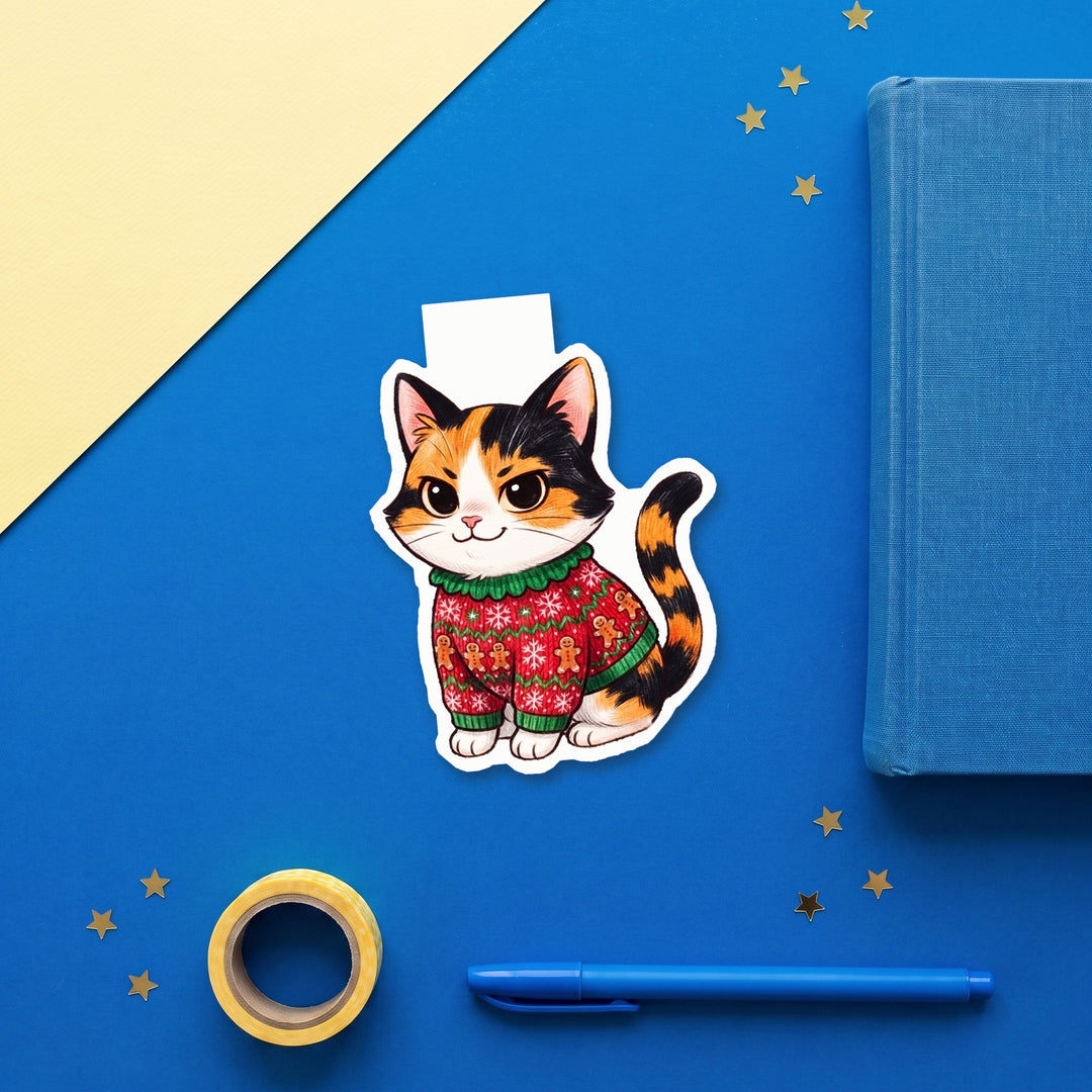 Tricolor calico cat in gingerbread ugly Christmas sweater kawaii magnetic bookmark flat lay with blue stationery – Designs by Mysh