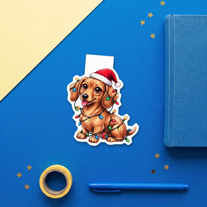 Red Dachshund in Santa hat tangled in Christmas lights kawaii magnetic bookmark flat lay with blue stationery – Designs by Mysh